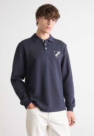 Polo à manches longues bleu marine avec col, trois boutons blancs et un logo de blason en blanc. Caractérisé par une coupe décontractée et un tissu doux.