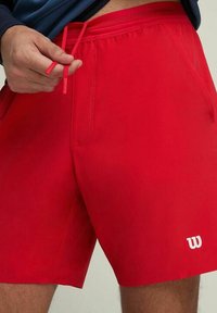 Hombre ajustando pantalones cortos rojos con cordón y logo de Wilson en el dobladillo, contra un fondo neutro.