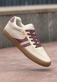 Beige sneaker with burgundy accents, airborne above a textured surface. Features include laces, logo, and contrasting sole color.