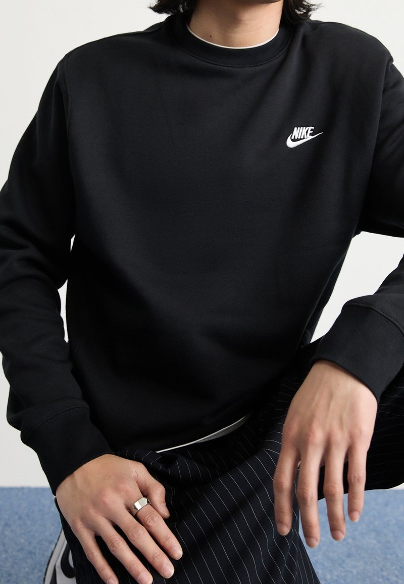 Sudadera de algodón negra con cuello redondo, que presenta un logo blanco de Nike en el pecho. Corte holgado, con mangas largas y puños acanalados.