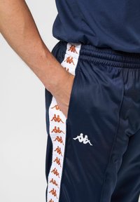 Marineblauwe sportbroek met witte zijkanten en een oranje logo patroon, en een klein wit logo op de linker dij. Soepele, lichte stof.