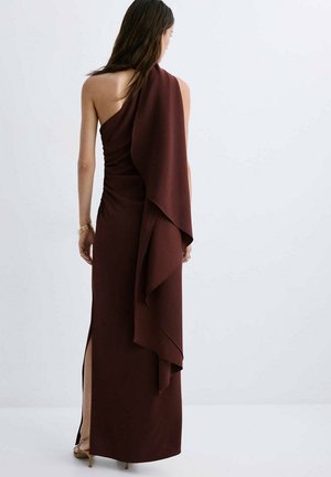 Femme portant une robe longue marron foncé asymétrique avec un détail de tissu drapé et une fente latérale, debout sur un fond clair uni.