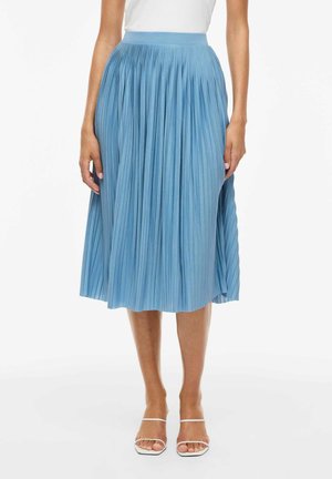 Kvinde iført en lyseblå plisseret midi-nederdel, hvid top og hvide åbne sandaler, stående mod en ensfarvet hvid baggrund.