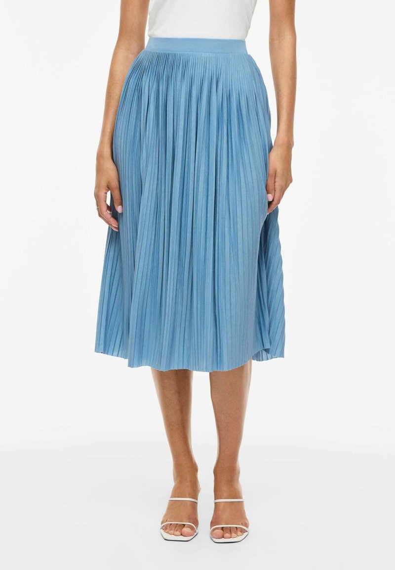 Kvinde iført en lyseblå plisseret midi-nederdel, hvid top og hvide åbne sandaler, stående mod en ensfarvet hvid baggrund.