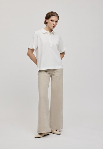 Chemise polo blanche en coton à manches courtes avec patte de boutonnage, associée à un pantalon large beige clair et des chaussures beiges à bouts pointus.
