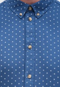 Sinine nööbitav triiksärk, millel on ühtlane tekstuur ja valgete täpiliste mustritega, terava kraega ning eespruugil beežid nupud.