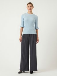 Femme debout portant un pull léger bleu ciel à côtes avec des manches à volants et un pantalon large foncé, ainsi que des chaussures à talons noires.