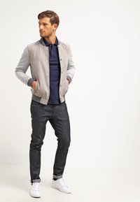 Siva bomber jakna s temno modro polo majico, temnimi kavbojkami in belimi supergami. Jakna ima gladko teksturo in gumbasto zapiranje.