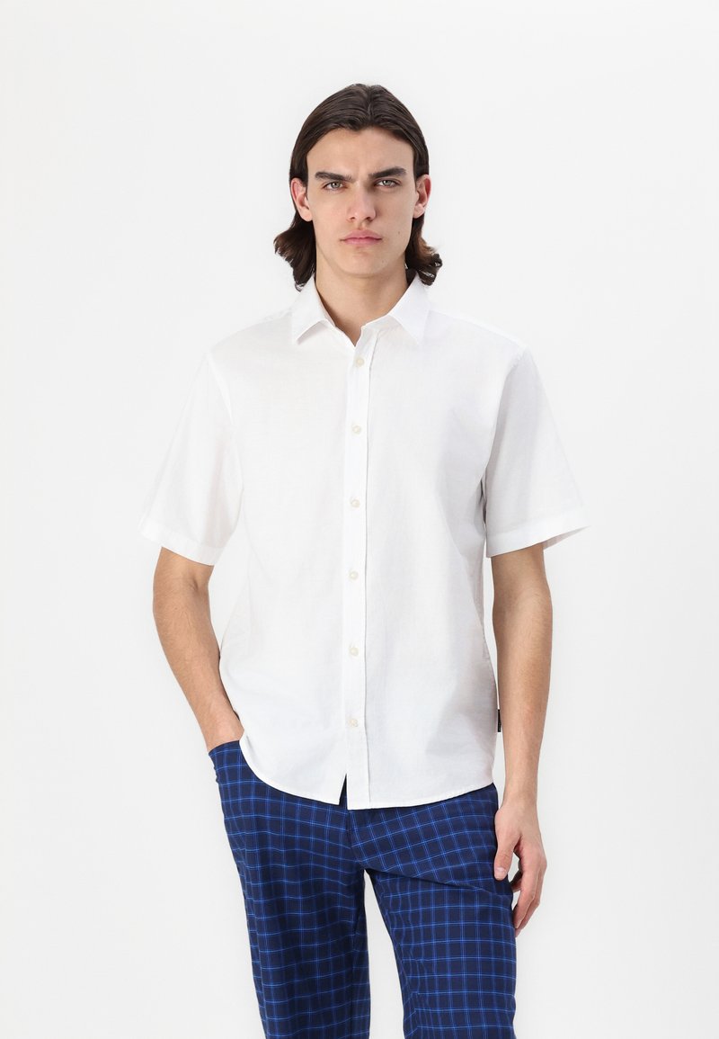 Camicetta bianca in lino a maniche corte con colletto button-down, con una texture liscia e una vestibilità comoda, abbinata a pantaloni a quadri blu.