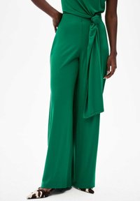 Groene wijde broek van gladde stof, met een gestrikte ceintuur in de taille en een vloeiende uitstraling, gecombineerd met schoenen met dierenprint.