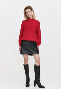 Czerwona dzianinowa sweter z wysokim kołnierzem, w połączeniu z czarną skórzaną mini spódnicą i czarnymi kozakami na wysokim obcasie do kolan. Gładka tekstura i zorganizowany styl.