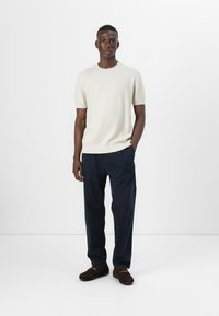Licht beige korte mouw trui gecombineerd met marineblauwe broek. De outfit wordt compleet gemaakt met bruin instap schoenen, tegen een eentonige witte achtergrond.