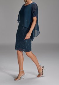 Femme portant une jupe en dentelle bleu marine avec des sequins et un haut cape transparent assorti, associée à des sandales à talons hauts argentées à brides.