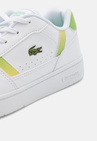 Sneaker in pelle bianca con accenti verde chiaro e giallo, perforazioni e logo Lacoste. Suola piatta con grip testurizzato.