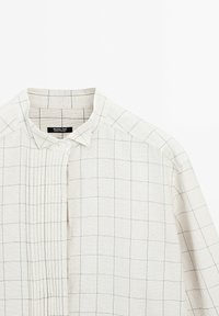 Chemise beige clair avec un motif en grille, dotée d'un col boutonné et de détails plissés. Fabriquée en tissu texturé avec des accents bleus.