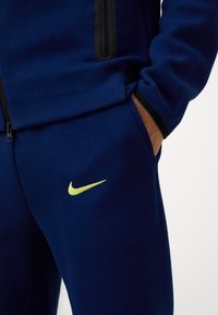 Marineblå Nike træningsdragt med et fremtrædende gult logo og lynlåslommer. Stoffet ser glat og strækbart ud, hvilket sikrer komfort og bevægelsesfrihed.
