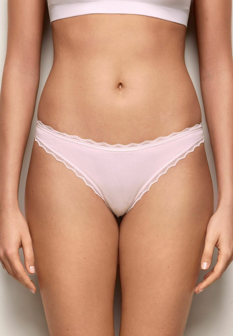 Mujer con ropa interior estilo bikini de encaje rosa pálido y top blanco corto, de pie con los brazos ligeramente levantados junto al cuerpo.