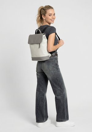 Naeratav noor naine, kes kannab musta t-särki, pleekinud musti teksapükse, valgeid tossusid ja halli-valget seljakotti, seisab ühevärvilise tausta ees.