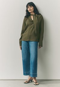 Olive green blouse with a draped neckline and tie detail, paired with wide-leg blue denim jeans with a hemmed finish and black sandals.