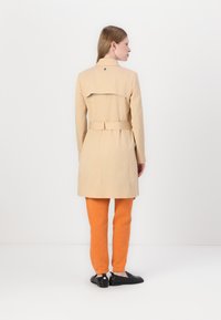 Manteau beige avec un détail de rabat à l'arrière, taille ceinturée et manches longues, associé à un pantalon orange vif et des chaussures noires.