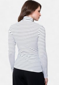 Dugi rukav, majica s lažnim turtleneck izradom u bijeloj boji s crnim vertikalnim crtama. Rebrasta tekstura, prilagođeni kroj i ravni šavovi. Model okrenut leđima.
