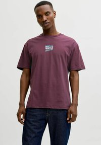 Vinrød bomulds-T-shirt med korte ærmer, med et grafisk tryk "WHISTLER" og "Blackcomb" på brystet. Båret med mørke jeans.