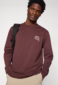 T-shirt de manga longa cor bordô, feito de tecido suave, com um pequeno gráfico branco no lado esquerdo do peito e com decote redondo.