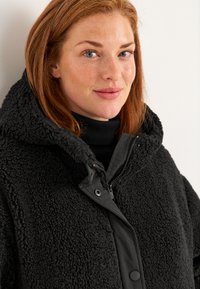 Zwarte, hooded jas met een gestructureerde, fuzzy buitenkant en een gladde voorpaneel. Voorzien van drukknoop- en ritssluitingen, gedragen over een zwarte coltrui.