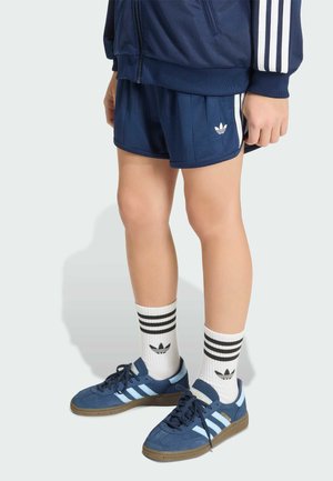 Kind draagt marineblauwe Adidas-short, bijpassend jasje, witte Adidas-sokken en blauwe Adidas-sneakers, staande tegen een lichte achtergrond.