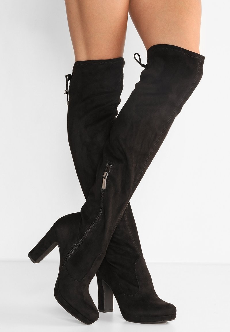 Bottes montantes en daim noir avec talons carrés et fermetures éclair latérales, portées sur des jambes croisées sur un fond blanc.