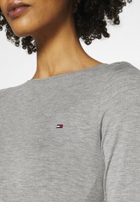 Grijs langemouwen T-shirt van zacht materiaal, met een ronde hals en een klein geborduurd logo in rood, wit en blauw op de borst.