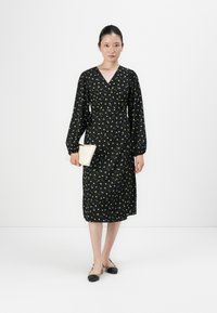 Woman wearing a black floral long-sleeve midi wrap dress, beige and black slingback shoes, holding a white clutch, standing against a plain background.
