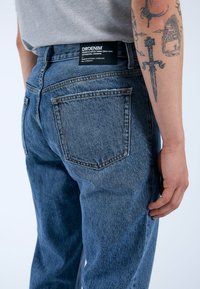 Jeans de mezclilla azul con un corte relajado, que presentan un parche trasero de cuero negro con logotipo, cinco bolsillos y costuras visibles.
