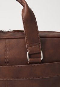 Brown leather bag with a smooth texture, featuring a flat handle and metal hardware. Stitched detailing along the sides.