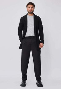 Homme debout les mains dans les poches, portant une chemise gris clair, un long cardigan noir, un pantalon noir et des chaussures noires, sur un fond uni.