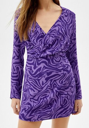Woman wearing a purple long-sleeve mini dress with dark purple abstract zebra stripes and a V-neck wrap bodice.