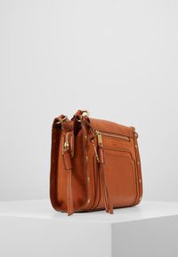 Brun læder crossover taske med guldfarvet hardware, lynlåslukning, dekorative nitter og flad design. Har vævet skulderrem og sidelomme.