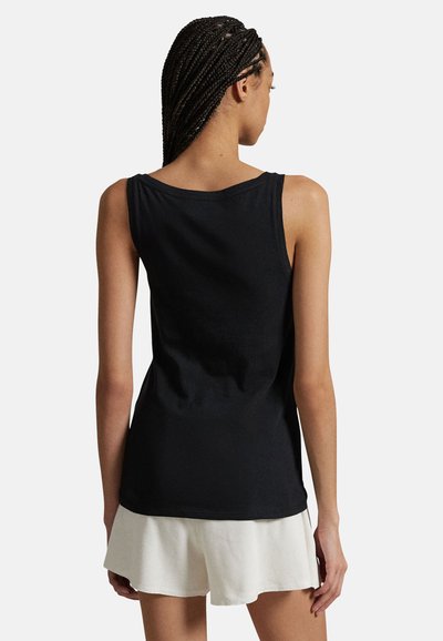 Femme aux cheveux tressés portant un haut noir sans manches et un short blanc texturé, debout de dos sur un fond uni.