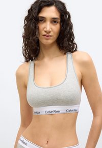 Γυναίκα με σγουρά σκούρα μαλλιά που φοράει γκρι αθλητικό σουτιέν και εσώρουχο με λευκές ελαστικές ταινίες με την επωνυμία Calvin Klein, ποζάροντας μπροστά σε λευκό φόντο.