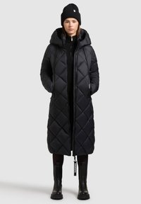 Čierny dlhý kabát v prevedení puffer s diamantovým prešívaním, prehnutou kapucňou a plným zipsom; skombinovaný s čiernymi čižmami po kolená.