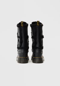 Botas de cuero negro hasta el tobillo con forro de felpa, cuello forrado y etiqueta de marca amarilla. Presentan suelas de goma gruesa y texturizada, y correas decorativas.