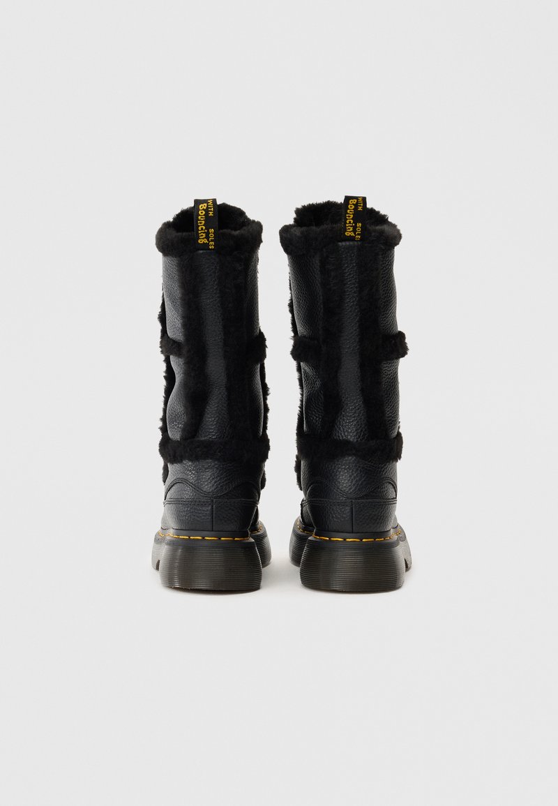 Bottines noires en cuir avec doublure en peluche, col rembourré et étiquette de marque jaune. Elles possèdent des semelles en caoutchouc texturé et chunky ainsi que des sangles décoratives.