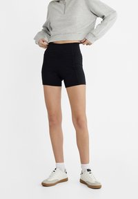 Svart hög midja shorts tillverkade av elastisk tyg, med en sömlös design och dekorativa sömnadsdetaljer. Bärs med en grå sweatshirt och sneakers.