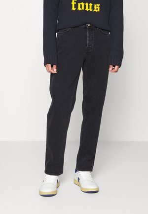 Osoba ubrana w czarne dżinsy, białe tenisówki z niebieskimi akcentami oraz ciemny sweter z żółtym napisem na jednolitym tle.