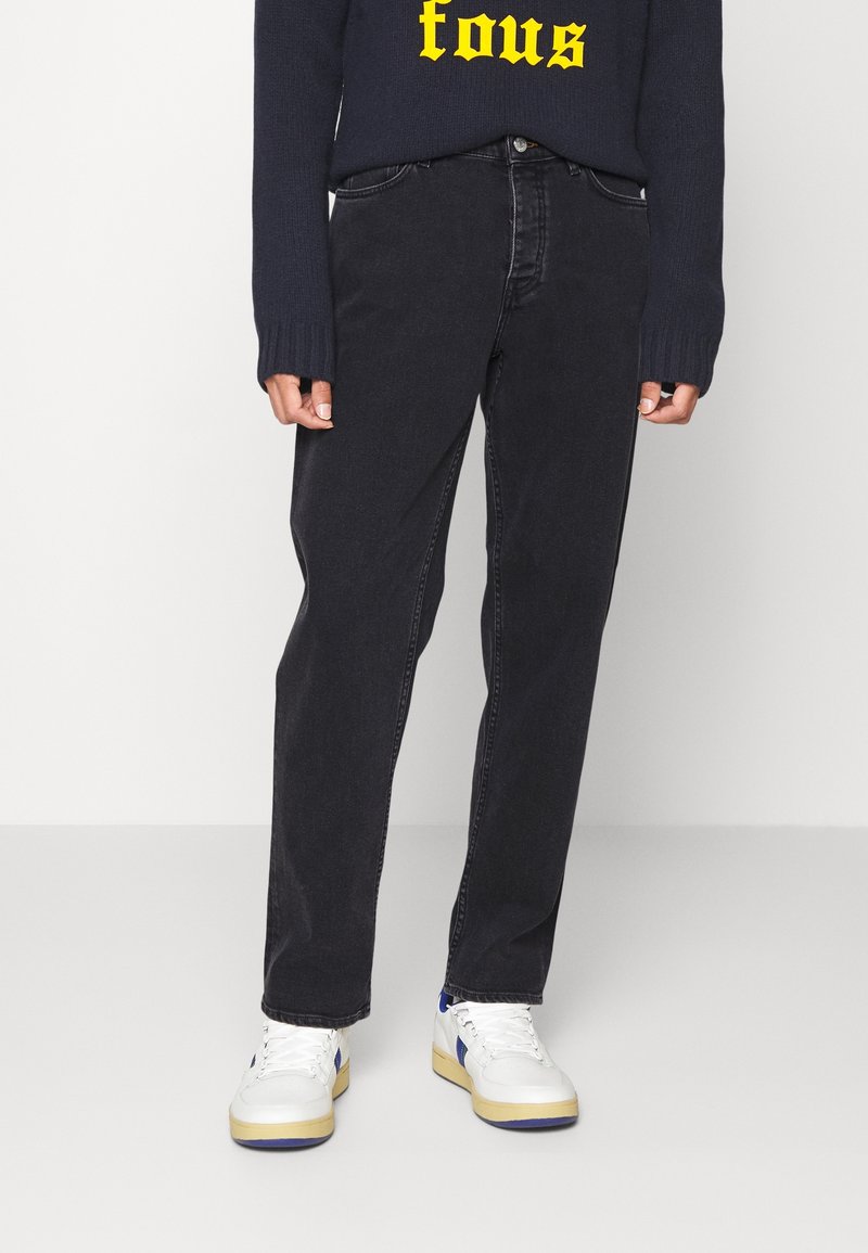 Personne portant un jean noir, des baskets blanches avec des touches bleues et un pull foncé avec un texte jaune sur un fond uni.