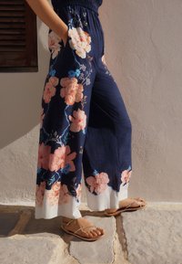 Pantaloni largi albăstri cu modele florale în nuanțe de piersic și albastru, având o tiv alb, asortați cu papuci tip flip-flop de culoare cămilă pe o suprafață de piatră.