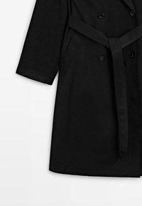Trench-coat noir à manches longues, boutons croisés, ceinture à nouer à la taille et poches latérales sur fond blanc.