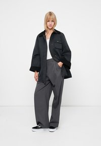 Giacca nera con colletto e due tasche frontali, abbinata a pantaloni grigi plissettati e sneakers nere. Il tessuto appare liscio e leggero.