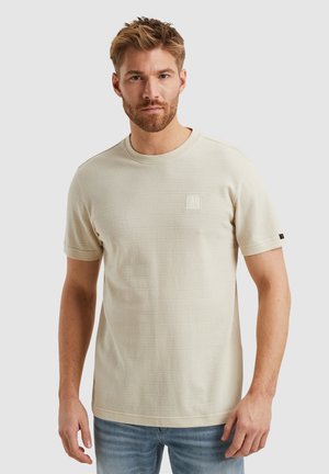 Lichtbeige getextureerde T-shirt met korte mouwen, een ronde halslijn en een klein vierkant logo op de borst. Gecombineerd met blauwe jeans.