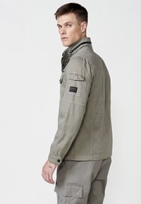 Khaki bunda so zipsovým golierom, gombíkovými vreckami a vyšitým logom na rukáve. Tkanina má hladkú, ľahkú textúru.